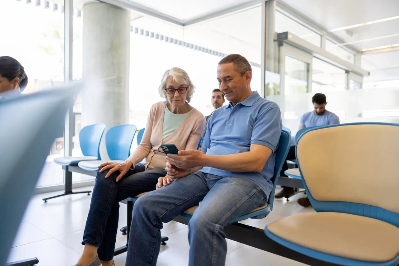 A patient and companion in a clinic waiting room consulting information on a mobile phone.